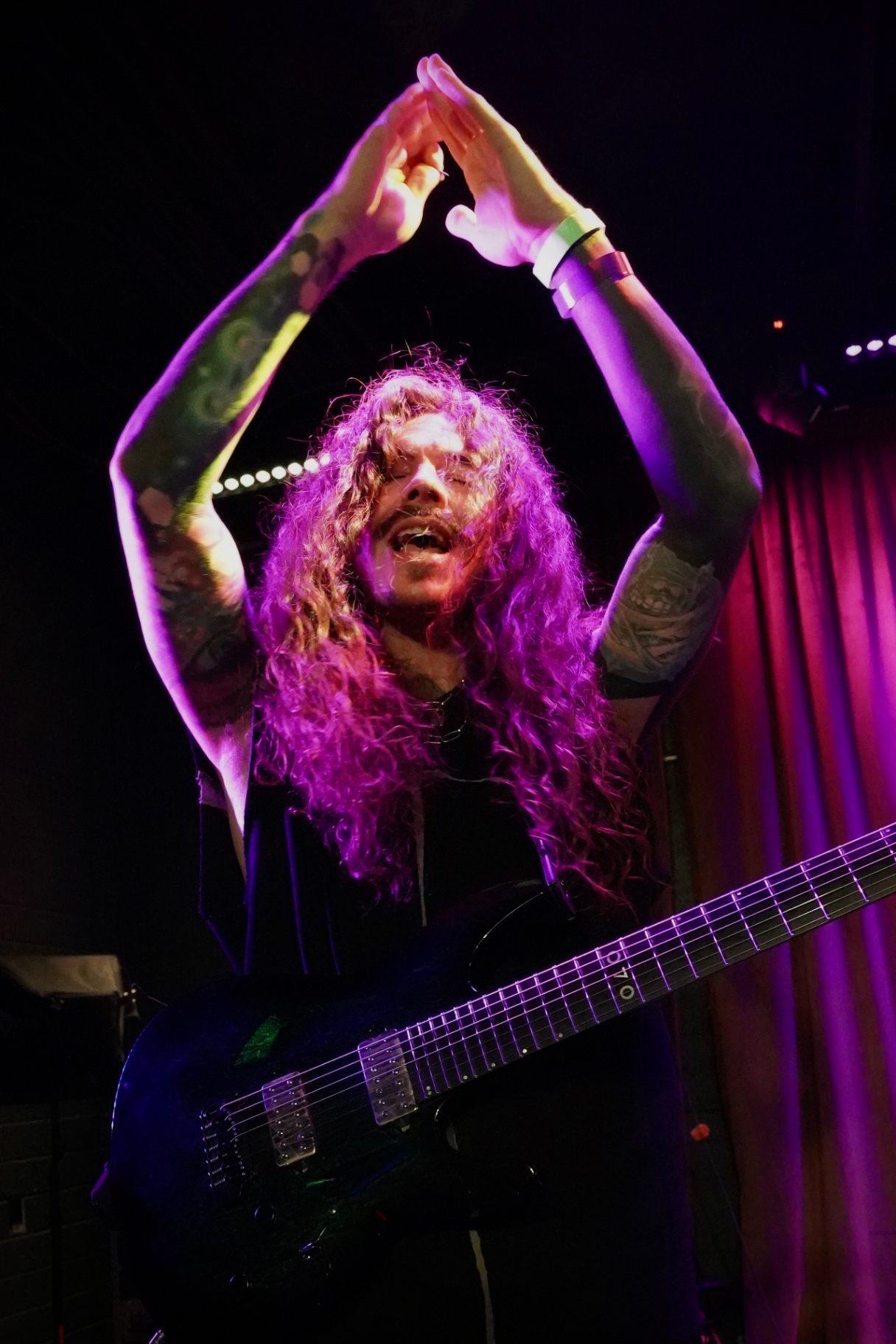 Curly-haired bassist raising hands overhead under purple light