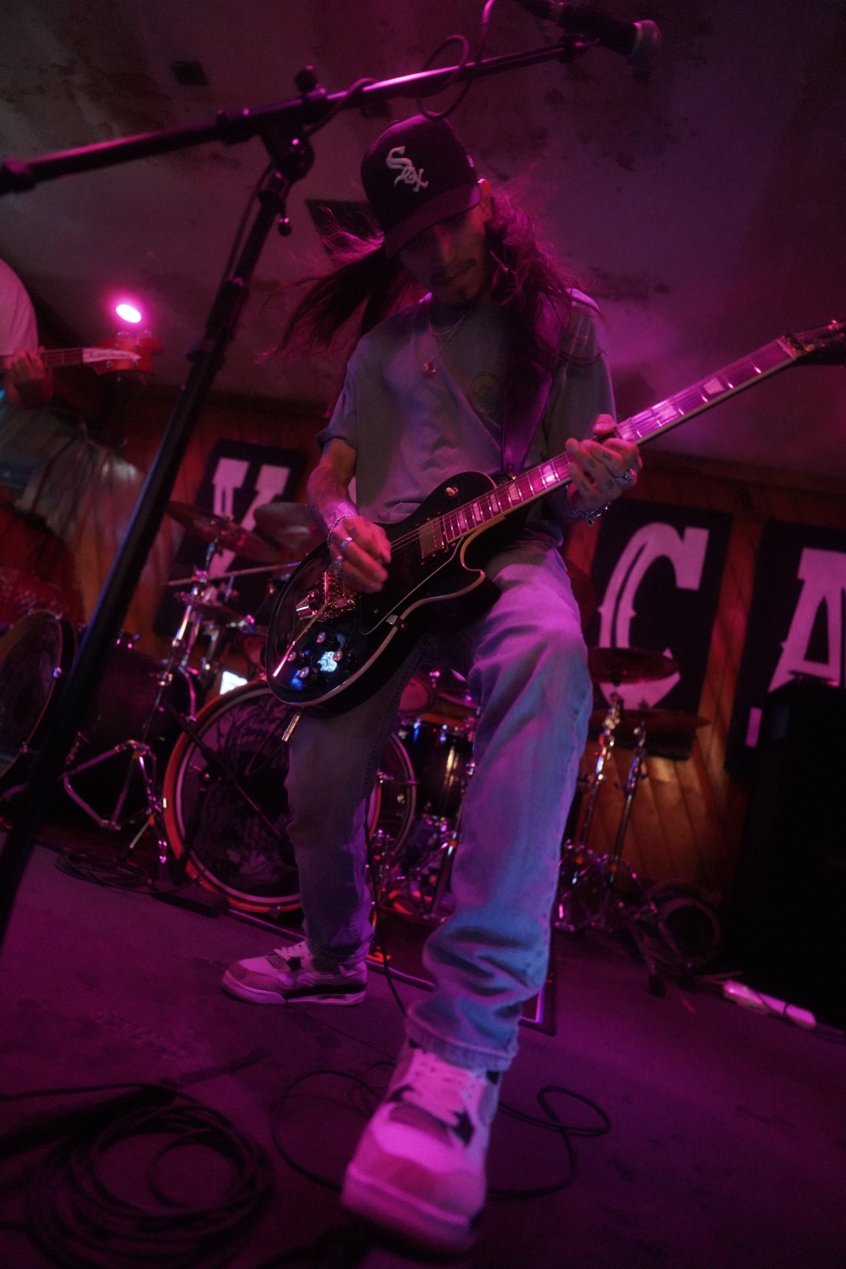 Long-haired guitarist mid-riff on a black Les Paul under magenta light