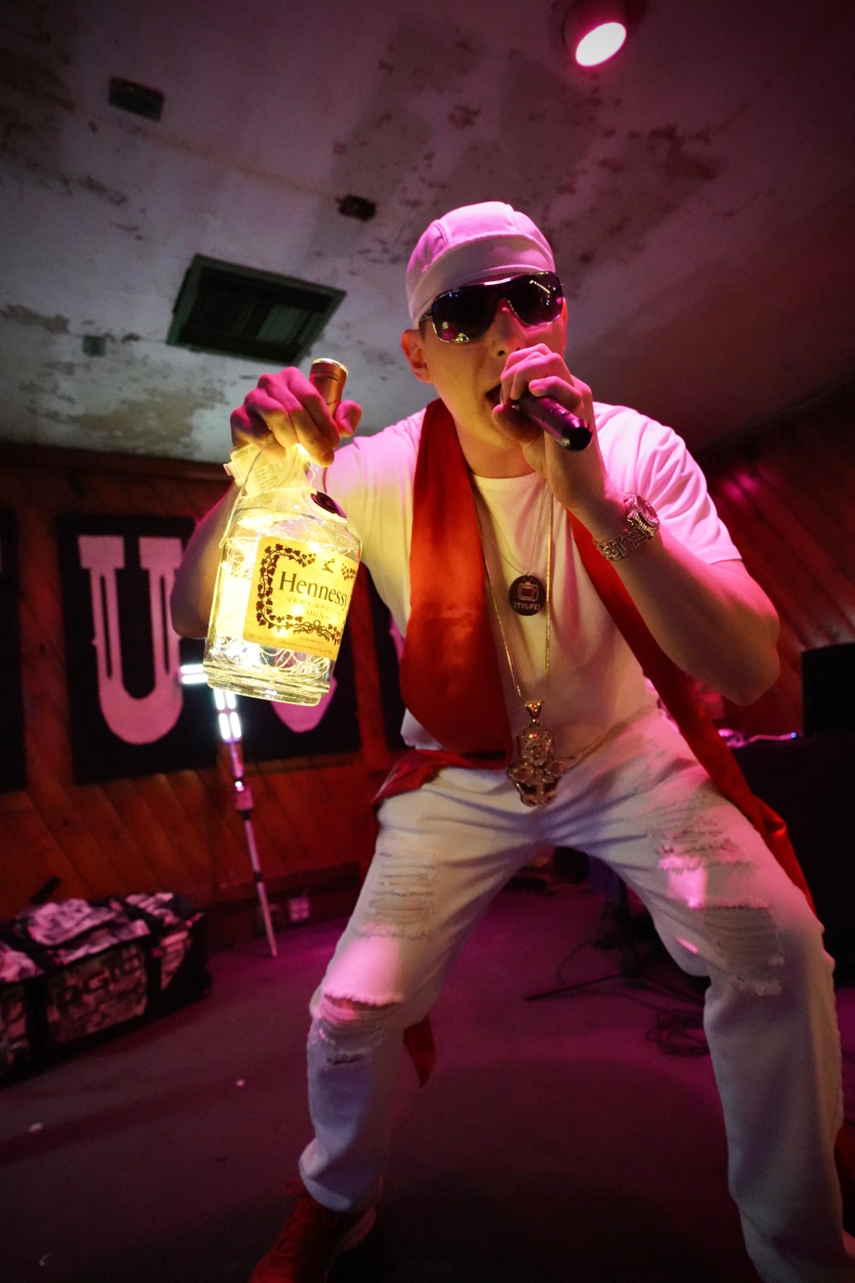 Rapper in all white holding a Hennessy bottle and mic under pink light